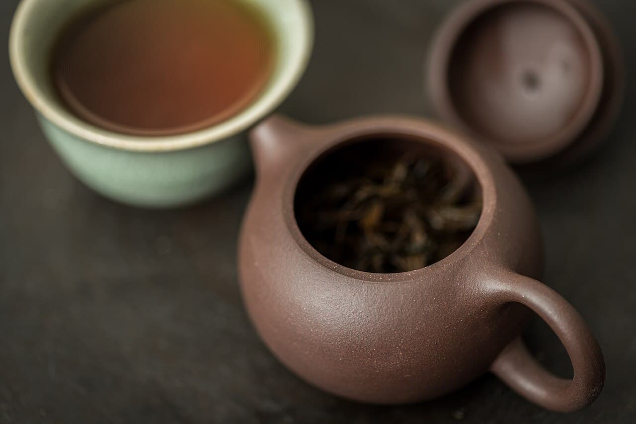 An open Yixing clay teapot holding tea leaves beside a cup of brewed tea and the pot's lid.