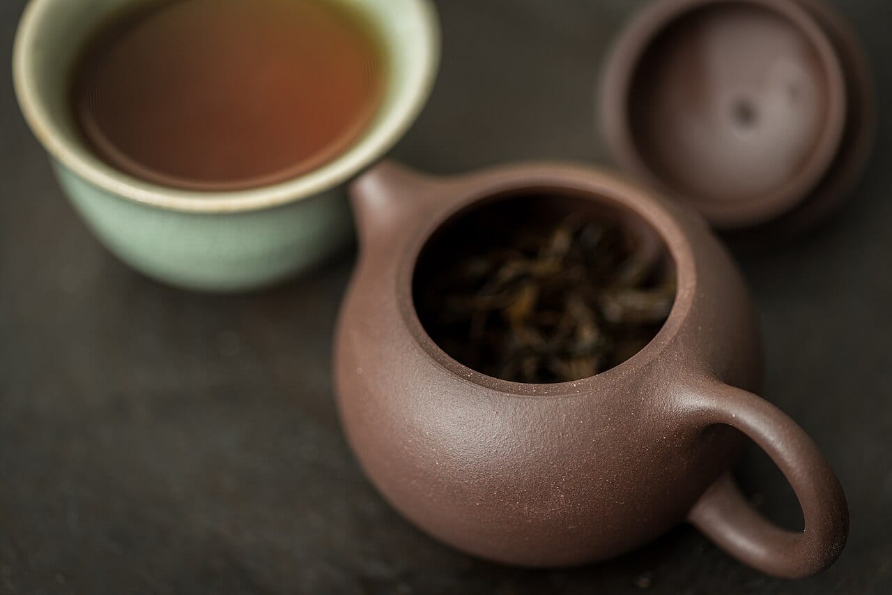 An open Yixing clay teapot holding tea leaves beside a cup of brewed tea and the pot's lid.