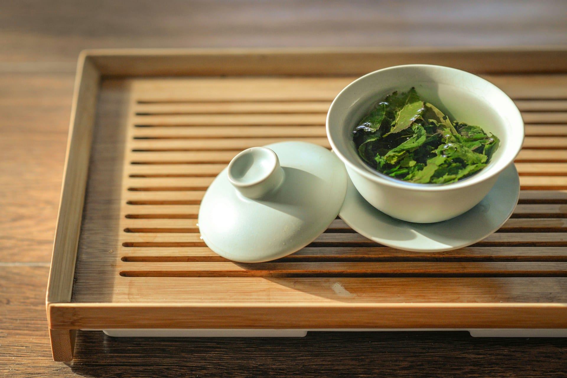 A white porcelain gaiwan of green tea, its lid resting beside it, on a bamboo tea tray in warm daylight